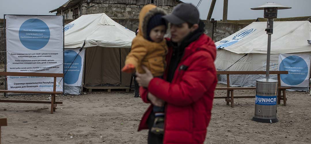 A young man holds his nephew as they walk past a UNICEF UNHCR Blue Dot centre